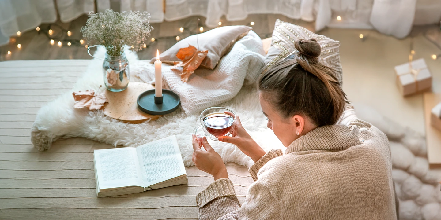 Femme en pleine ambiance cocooning lisant un livre et tenant une tasse de thé, symbole de rituels d’hiver et de recentrage spirituel.